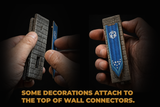 Two hands holding miniature wall decorations against a black background. The left hand holds a gray stone-textured rectangular piece, and the right hand holds a blue banner with a white symbol mounted on a gray stone-textured piece.