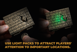 Two hands hold miniature stone-textured board game pieces against a black background, with one piece lit green by a light pack. Gold text at the bottom reads, 'USE LIGHT PACKS TO ATTRACT PLAYERS' ATTENTION TO IMPORTANT LOCATIONS.